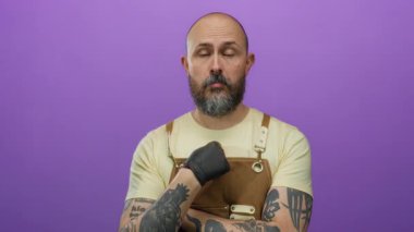 Önlüklü ve siyah eldivenli yakışıklı sakallı adam mor bir arka plana karşı duruyor, düşünceli bir ifade sergiliyor..