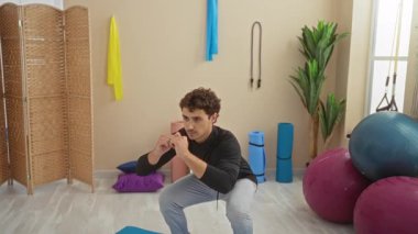 Spor salonunda spor aletleriyle egzersiz yapan genç bir adam. Doğal ışık ve bitkilerle çevrili renkli egzersiz topları ve yoga minderleri de dahil..