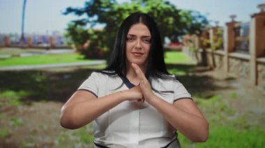 Park ortamında kıvrımlı, el kol hareketleri yapan, beyaz gömlek giyen, siyah saçlı, yeşilliklerle çevrili, açık havada doğal ışıkla iletişim kuran bir kadın..