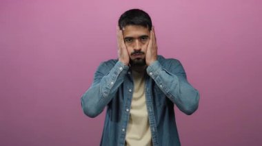 Genç İspanyol bir adam, elleri yüzünde stresli görünüyor, pembe izole edilmiş bir arka planda kot gömlek giyiyor..