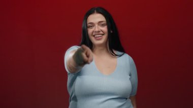Gülümseyen ve parlak kırmızı arka plana işaret eden bir kadın, özgüveni ve pozitifliği somutlaştıran, bir stüdyoda geçen, açık mavi bir bluzla artı boy temsil eden bir kadın..