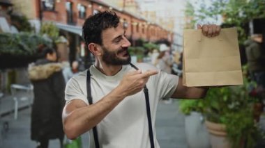 Sakallı İspanyol bir adam yeşillik ve şehir zeminiyle çevrili açık hava sahnesinde kahverengi kesekağıdına sevinçle işaret ediyor..