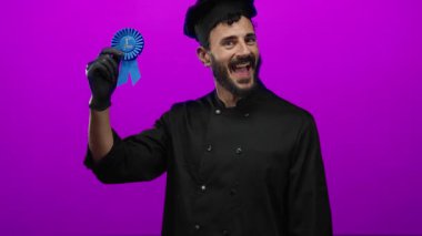 Siyah şef üniformalı İspanyol adam, mor arka plana karşı birinci sınıf mavi kurdeleyle kutlama yapıyor, neşe ve başarıyı ifade ediyor..