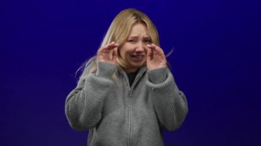 Gri kazaklı bir kadın mavi bir arkaplanda korkmuş görünüyor, korku ve endişe duygularını ifade ediyor, sarı saçlı ve izole bir çevrede, bir huzursuzluk duygusu somutlaştırıyor.