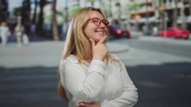 Açık havada gülümseyen sarı saçlı, gözlüklü ve beyaz gömlekli bir kadın işlek bir şehir caddesinde bulanık bir arka plana karşı düşünceli bir ifadeyle duruyor..