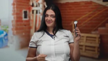 Bir inşaat alanında elinde makyaj fırçasıyla elinde tuğladan bir arka planla gülümseyen bir kadın güzellik ve inşaat temalarının bir karışımını sergiliyor..
