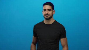 Genç İspanyol bir adam parlak mavi bir duvara karşı düşünceli bir şekilde el kol hareketi yapar, kendinden emin ve çekici bir tavır sergiler..