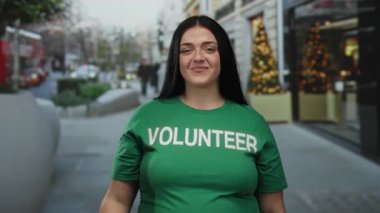 Yeşil gönüllü gömlekli kıvrımlı genç bir kadın, gülümsüyor ve şehrin arka planında şenlikli süslemeler ve kırmızı bir otobüsle yukarı doğru işaret ediyor..