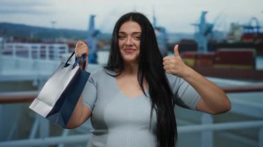 Alışveriş çantalı genç bir kadın bir yolcu gemisinde baş parmak hareketi yapıyor. Manzaralı bir liman zeminine karşı gösteriş yapıyor. Seyahat ve alışveriş havasını karıştırıyor..