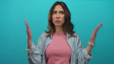 Şaşkın bakışlı bir kadın pembe gömlek ve kot ceket giyen, kafa karışıklığı ve merakı taşıyan canlı mavi bir arka plana el kaldırıyor..