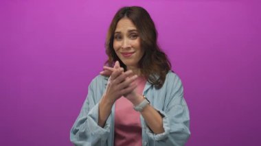 Pembe arka planda gülümseyen bir kadın, elleri sımsıkı bağlanmış, bir stüdyo ortamında neşe ve güven ifade eden, günlük mavi bir gömlek ve saat giyen, gençlik enerjisi sergileyen.