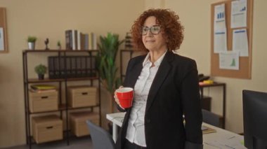 Ofiste elinde kırmızı bardakla duran, gözlüklü ve ceketli bir kadın. Etrafı raflar ve bitkilerle çevrili, modern bir iş yeri ortamında..