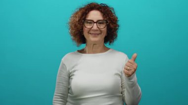 Gülümseyen, esmer, orta yaşlı, gözlüklü, soyutlanmış, mavi arkaplan, sıradan tarz, baş parmaklar yukarı, mutluluk, pozitif ifade.