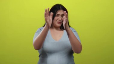 Kadın, elleri kaygılı bir şekilde parlak sarı arkaplana karşı yüzünü kapatıyor. Günlük mavi gömlekler giyiyor ve izole edilmiş duygu kavramını sergiliyor..