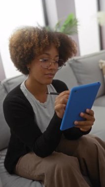 Oturma odasındaki rahat bir tablette yazan kadın, yumuşak yastıklarla çevrili, günlük kıyafetler ve gözlük takan, evde rahat bir anın tadını çıkaran..