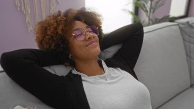 Kıvırcık saçlı ve gözlüklü, rahat, modern apartman dairesinde dinlenen ve boş vakitlerin tadını çıkaran bir kadın..