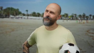 Güneşli sahilde futbol topu tutan kel sakallı adam mavi gökyüzünün altında palmiye ağaçlarıyla çevrili açık havanın tadını çıkarıyor..