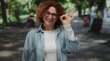 Yeşil ağaçlar ve açık hava ışıklarıyla çevrili, parkta gülümseyen bir kadın, huzurlu bir ortamda keyif ve güven anı yakalıyor..