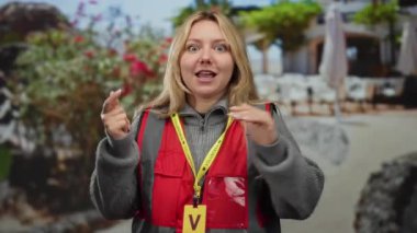 Kırmızı yelekli, gönüllü jestli bir kadın arka planda çiçekler olan bir şehirde terasta..