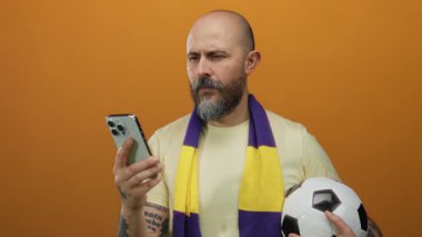 Sakallı bir adam akıllı telefon ve futbol topunu turuncu arka planda tutuyor. Eşarp takıyor. Dalgın görünüyor..
