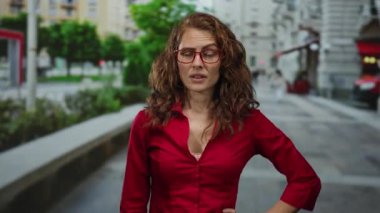 Kırmızı tişörtlü, kıvırcık saçlı ve gözlüklü bir kadın. Arka planda bulanık binalar ve yeşil ağaçlarla duygularını ifade ediyor..