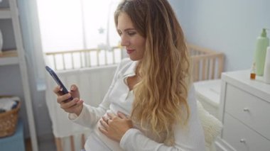 Akıllı telefonlu hamile bir kadın rahat bir yatak odasında beşiğin yanında oturuyor, karnını kucaklıyor ve mutlu bir şekilde gülümsüyor..