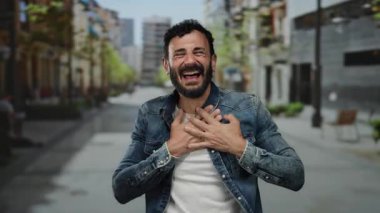 Sakallı İspanyol bir adam gülerek ve bir şehir sokağında, kot ceket giyip dışarıda mutluluğu ifade ederek neşeyle işaret ediyor..