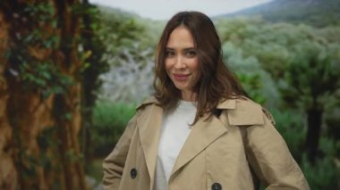 Açık gökyüzünün altında bej bir ceket giyen, açık havada gülümseyen bir kadın doğal manzara ve yeşillik arasında gevşemiş ve neşeli bir hava sergiliyor.