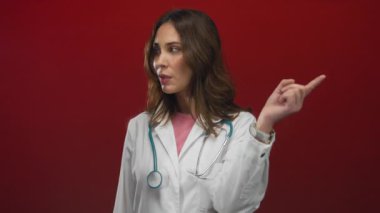 Beyaz önlüklü İspanyol bayan doktor ve steteskop kızgın bir şekilde kırmızı arka plana işaret ediyor, tıbbî ortamda hayal kırıklığı ve otoriteyi somutlaştırıyor..
