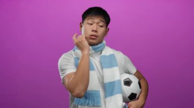 İzole edilmiş pembe bir arka planda futbol topunu tutan ve açık mavi eşarp takan genç adam ciddi bir ifadeyle işaret ediyor..