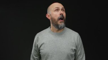 Sakallı bir adam, izole edilmiş siyah bir arkaplana şaşkınlıkla bakıyor gri bir süveter giyiyor. Hislerini minimalist ortamda ifade ediyor..