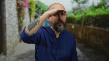 Sakallı ve dövmeli bir adam güneşli bir günde yeşil ve çiçekli mavi gömlekli bir caddede uzaklara bakıyor.