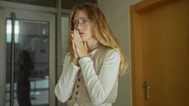 Kapalı elleriyle, sarı saçlarıyla ve gözlükleriyle içeride dua eden bir kadın, bir ofis odasında yansıyor, huzurlu bir ortamda umut ya da minnettarlığını dile getiriyor..