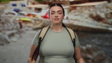 Sırt çantalı, resimli bir plajda duran kendinden emin genç bir kadın. Arka planda çarpıcı bir deniz manzarası olan jestin ortasında çekilmiş. Barışçıl bir açık hava deneyimi yaşıyor..