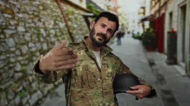 Askeri üniformalı İspanyol bir adam, sokak ortamında sakallı jestler yapıyor, bulanık bir şehir zemininin ortasında miğfer tutuyor, aciliyet ya da talep duygusunu iletiyor..