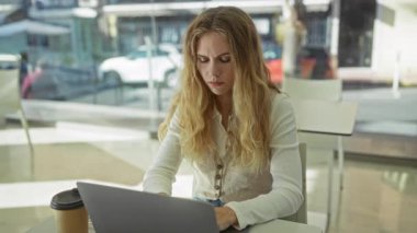 Modern ofiste dizüstü bilgisayarda çalışan bir kadın, kahve, odaklanmış, düşünceli, sarışın, beyaz bluzlu, masada oturan, iç mekan, kentsel ortam.