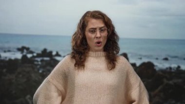 Gözlüklü kadın bulutlu bir gökyüzünün altında kayalık sahilde yüzünü buruşturuyor, rahat bir bej kazak giyiyor, açık havada serin okyanus esintisinin tadını çıkarıyor..