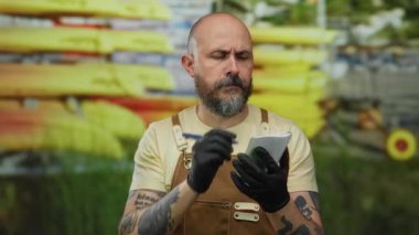 Sakallı, eldiven ve önlüklü bir adam arka planda kanoyla notlar yazar. Odak ve konsantrasyonu canlı manzara ve dinamik ortam arasında yakalar..