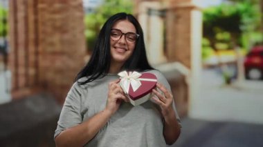Şehir caddesinde elinde kalp şeklinde bir hediye kutusu olan bir kadın neşeli bir ifadeyle, gözlük takıyor ve gündelik giysiler giyiyor, aydınlık bir günde şehir mimarisi ile çevrili..