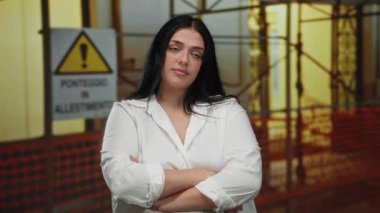 Arka planda İtalyan uyarı tabelasıyla inşaat alanında güvenle duran bir kadın, geniş bir moda tarzını açık havada sergiliyor..