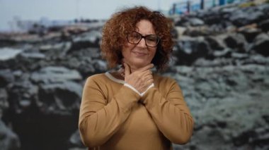 Gözlüklü bir kadın, kayaların ve okyanusun kenarında duruyor, kahverengi bir kazak giyiyor, düşünceli görünüyor, deniz kenarındaki sükuneti yansıtıyor, doğal güzellik ve sükuneti sergiliyor..