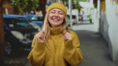 Sarı süveter ve bereyle gülen kadın şehir merkezinde gündüz vakti mutluluğunu ve neşesini ifade ediyor..