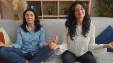 Orta yaşlı kadınlar, rahat bir oturma odasındaki kanepede birlikte meditasyon yapıyorlar. Huzur ve kardeşçe bir bağ kuruyorlar. Bitkiler ve yumuşak dekorla huzurlu bir ev ortamında..