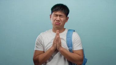 Asyalı kökenli genç adam mavi arka plana karşı umut dolu bir ifadeyle dua ediyor. Duygularını ve samimiyetini gösteriyor..