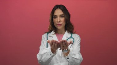 İspanyol kadın doktor kameraya doğru elini uzatıyor boynunda steteskopla profesyonel bir tıbbi bağlamda izole edilmiş bir kırmızı arka planda..