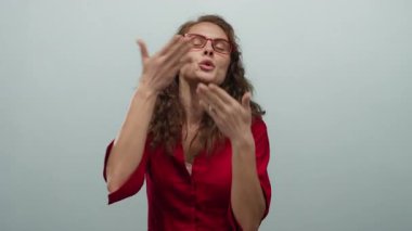 Kırmızı bluzlu, gözlüklü, öpüşen bir kadın izole edilmiş beyaz bir arka plan duvarına yaslanmış neşeli bir ifade ve vücut dili sergiliyor..