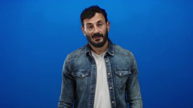 Sakallı, kot tişörtlü ve beyaz tişörtlü İspanyol bir adam mavi arka planda belirgin jestler yapıyor..