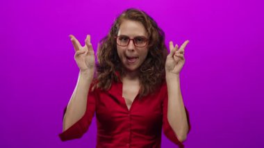 Kırmızı tişörtlü, gözlüklü bir kadın, parlak pembe arka planda parmakları olan tırnak işaretleri kullanıyor. İğneleme ve odaklanmış ifade ile alay ediyor..