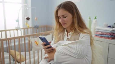 Yatak odasında akıllı telefon kullanan bir kadın battaniyeye sarılı bebeği tutuyor. Yanında beşik var..