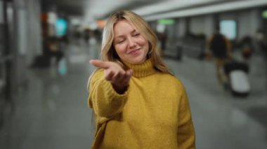 Havaalanı terminalinde gülümseyen sarı süveter giyen, el uzatılmış sarı saçlı, kapalı mekan ortamında hoş geldin jesti yapan bulanık arka planlı bir kadın.